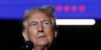 A man in a suit speaking into a microphone with a serious expression