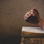 Hands clasped in prayer over an open book on a wooden table