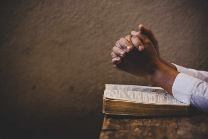 Hands clasped in prayer over an open book on a wooden table