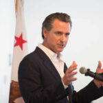 Water Crisis Politics: Is Newsom’s Plan Enough? A man in a suit speaking into a microphone with a California flag in the background