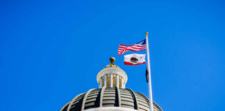 View of a state capitol dome with American and California flags against a blue sky