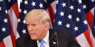 A man in a suit speaking at a podium with American flags in the background