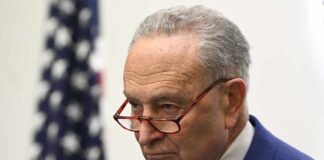 A serious-looking man in a blue suit with glasses, standing near an American flag