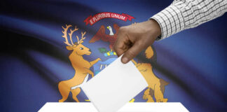 Hand placing a ballot into a ballot box with the Michigan state flag in the background
