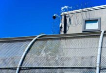 Security camera mounted above a prison fence with barbed wire against a blue sky