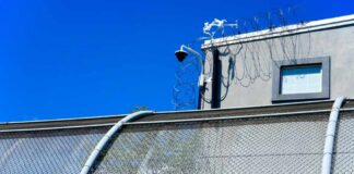 Security camera mounted above a prison fence with barbed wire against a blue sky