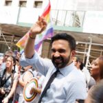 Smiling man waving at a pride parade with a rainbow flag in the background