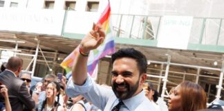 Smiling man waving at a pride parade with a rainbow flag in the background