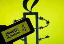 Amnesty International’s SHOCKING Accusations EXPOSED A hand holding a mobile device displaying the Amnesty International logo against a yellow background