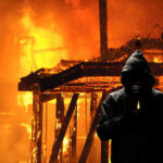 A hooded figure holding a lighter in front of a burning structure
