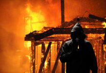 A hooded figure holding a lighter in front of a burning structure