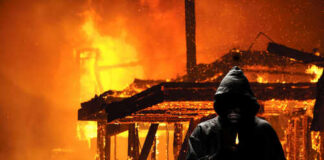 A hooded figure holding a lighter in front of a burning structure