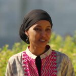 A woman in traditional attire smiling outdoors with greenery in the background