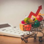 A shopping cart filled with colorful grocery items next to a calculator and an upward trend arrow