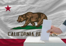 A hand placing a ballot into a box in front of the California state flag