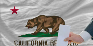 A hand placing a ballot into a box in front of the California state flag