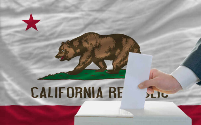 A hand placing a ballot into a box in front of the California state flag