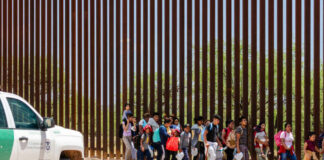 Drones of Terror: Cartels Clear Out Communities Group of families and children walking near a border wall with a customs vehicle nearby