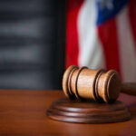 Judicial Misconduct COVER-UP—Dark Secrets Inside Courts A wooden gavel resting on a round base with a blurred American flag in the background