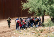 A group of migrants gathered near a border wall with a border patrol agent nearby