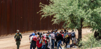 A group of migrants gathered near a border wall with a border patrol agent nearby
