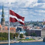 Latvian flag flying over a cityscape with church spire and river