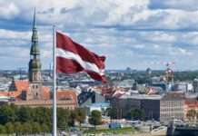 Latvian flag flying over a cityscape with church spire and river