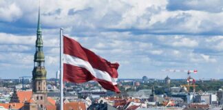Latvian flag flying over a cityscape with church spire and river