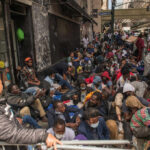A large group of people sitting on a city street, some wearing masks, in a crowded urban setting