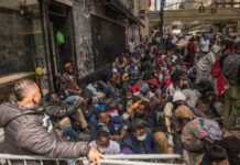 Mystery Attack Shakes Iran A large group of people sitting on a city street, some wearing masks, in a crowded urban setting