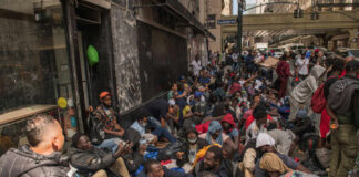 A large group of people sitting on a city street, some wearing masks, in a crowded urban setting
