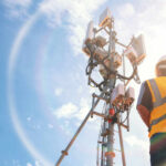 Telecommunication worker in safety gear standing near a cell tower under a blue sky