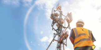 Telecommunication worker in safety gear standing near a cell tower under a blue sky