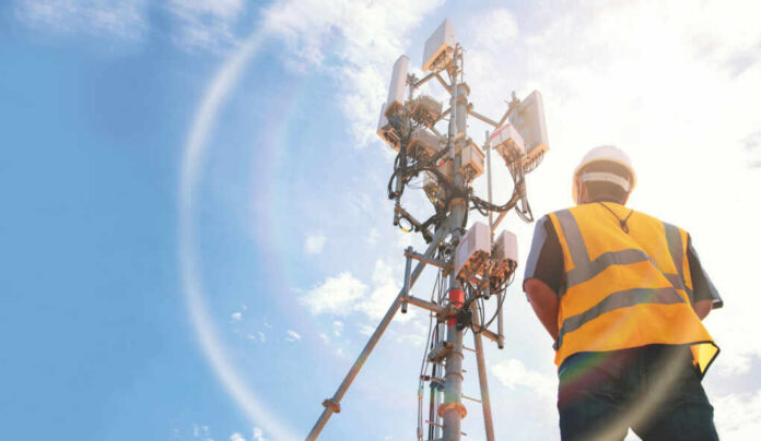 shutterstock_2106490226 Telecommunication worker in safety gear standing near a cell tower under a blue sky