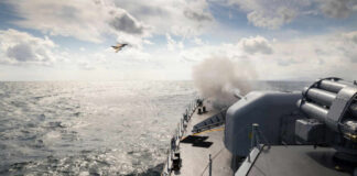 Power Play At Sea—Outcome Hidden, Risks Huge Naval ship firing a cannon with a fighter jet flying overhead