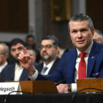 A man in a blue suit speaking at a senate hearing with a panel of officials behind him