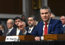 Pentagon Chief Torches Europe’s Borders A man in a blue suit speaking at a senate hearing with a panel of officials behind him