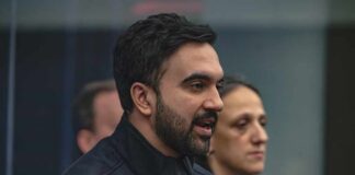 A man in a mayor's jacket speaking at a press conference with a woman in the background