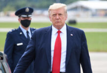 A political figure in a suit with a red tie walking outdoors, accompanied by a masked military officer