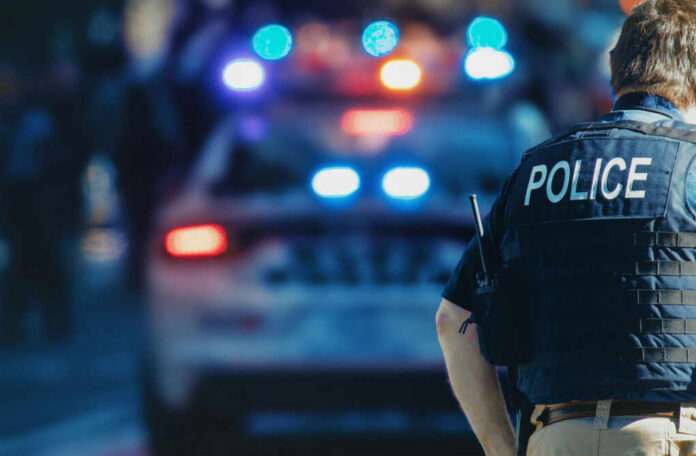 shutterstock_1543591088 Police officer in a tactical vest standing in front of a police car with flashing lights