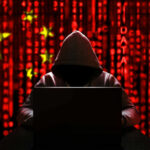 A hooded figure sitting in front of a laptop with a red digital background