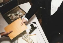 Two people exchange a manila envelope over a table with cash and a handgun