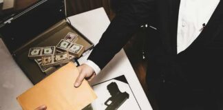 Two people exchange a manila envelope over a table with cash and a handgun