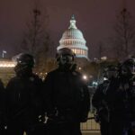 Riot police in protective gear stand before a domed capitol building at night
