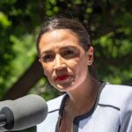 Dems Bench AOC In Swing Seats woman speaking at an outdoor microphone