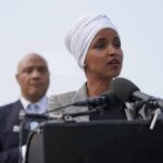 Marriage Fraud Bombshell Shadows Ilhan Omar Woman in white headwrap speaking at microphones outdoors