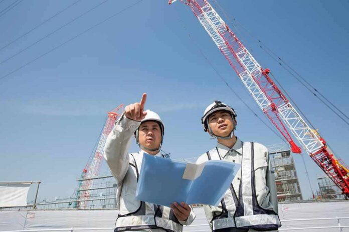 Two construction workers reviewing plans at a site with cranes