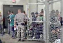 Murderer Vanishes – Fence Cam Stuns Guards People standing inside chain-link holding area with an officer nearby