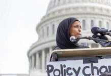 Grand Jury Targets Omar’s Inner Circle Ilhan Omar speaking at a podium with microphones in front of the U.S. Capitol