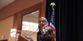 Omar’s Alias Bombshell Engulfs Congress Ilhan Omar speaking at a podium with a microphone